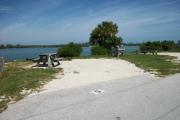 The back has a view of the inlet. The front is open to the street. There is a gravel drive in the center of the site. There is a picnic table on the left side with a fire ring behind it. A grassy area is on the right.