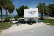 The back has a view of the inlet. The front is open to the street. There is a gravel drive in the center of the site. Sable palms are on both sides of the site. A picnic table is on the left. A white RV is in the driveway.