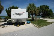 The back has a view of the inlet. The front is open to the street. There is a gravel drive in the center of the site. Palm trees are on the right. A picnic table is on the left. A white RV sits in the driveway.