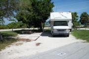 The back has a view of the inlet. The front is open to the street. There is a gravel drive in the center of the site. There is a picnic table under a large cedar on the left. A white RV sits in the driveway. A grassy area is on the right.