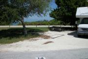 The back has a view of the inlet. The front is open to the street. There is a gravel drive in the center of the site. A tree shades the drive next to a white RV.  A picnic table sits under a large cedar on the left.