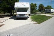 The back has a view of the inlet. The front is open to the street. There is a gravel drive in the center. A white RV sits in the driveway. An open grassy area is on the right. A picnic table under a large cedar is on the left.