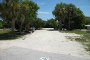 The front is open to the street and the back faces a street. There is a gravel drive in the center. A picnic table sits on the left. There is an open grassy area with trees in the back corner on the right. Trees line the driveway on the left.