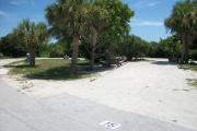 The front is open to the street and the back faces a street. There is a gravel drive in the center of the site. A street wraps around a grassy area on the left side of the site. Trees that line the driveway shade a picnic table on the left.