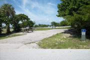 The campsite is handicap accessible. The front is open to the street. The back faces a street. There is a gravel drive in the center of the site. Both sides have a variety of trees. The left side has a picnic table.