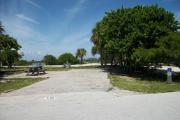 The campsite is handicap accessible. The front is open to the street and the back faces a street. There is a gravel drive in the center of the site.  Both sides of the site have several trees. The left side of the site has a picnic table next to a fire ring.