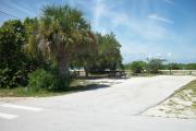 The campsite is handicap accessible. The front is open to the street and the back faces a street. There is a gravel drive in the center of the site. The left side has a picnic table next to trees with a fire ring behind it.