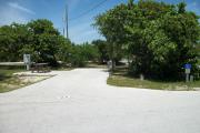 The campsite is handicap accessible. The front is open to the street. The back faces a street. There is a gravel drive in the center of the site. A picnic table is on the left. The right has a large sea grape.