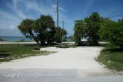 The front is open to the street and the back faces a street. There is a gravel drive in the center of the site. There are palm trees next to fire ring at the far left corner of the site. A picnic table is at the back. Sea grapes are in the right corner.
