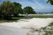The front is open to the street and the back faces a street. There is a gravel drive in the center. There is a picnic table with a fire ring behind it next to sea grapes and palms on the left.