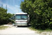 The front is open to the street and the back faces a street. There is a gravel drive in the center of the site. There is a large white RV in the driveway. A picnic table is on the left. Trees surround the site.
