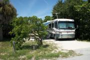 The front is open to the street and the back faces a street. There is a grave drive in the center of the site. A picnic table is on the left. A white RV sits in the driveway. Trees surround the site.