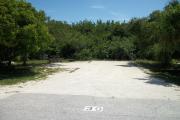 The front is open to the street. The back is filled with foliage. There is a gravel drive in the center of the site. The right side has several trees. There is a picnic table next to a fire ring on the left.