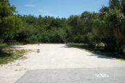 The front is open to the street. The back is filled with foliage. There is a gravel drive in the center of the site. The left side has a picnic table next to a fire ring. Trees surround the site.