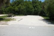 The front is open to the street. The back is filled with greenery. There is a gravel drive in the center of the site. A picnic table next to a fire ring is on the left. Trees are on the right.