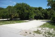 The front is open to the street. The back is filled with greenery. There is a gravel drive in the center of the site. A picnic table is shaded by large cedars and sea grapes on the left.