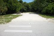 The front is open to the street.  The back is filled with greenery. There is a gravel drive in the center of the site. The left side has a crosswalk in front of it. A fire ring is behind a picnic table on the left.  Trees surround the site.